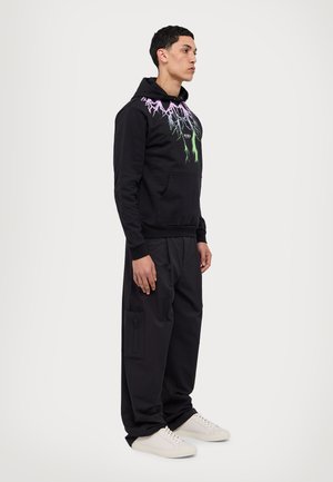 Felpa nera con grafica di fulmini vivaci, dotata di tasca a marsupio e polsini a coste. Abbinata a pantaloni neri larghi e sneakers chiare.