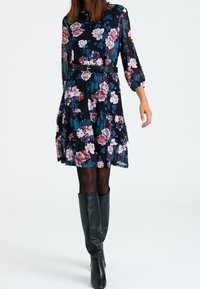 Robe en chiffon à fleurs avec un fond bleu foncé, manches longues, ourlet à volants et ceinture noire. Portée avec des bottes noires montantes jusqu'aux genoux.