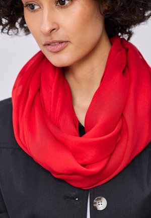 Kiharatukkainen nainen, jolla on kirkkaanpunainen huivi kaulassaan ja tumma napitettu takki.