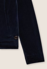 Felpa in velluto a coste blu navy con tasca laterale e piccolo logo circolare in hardware dorato. Texture morbida con polsini a coste.