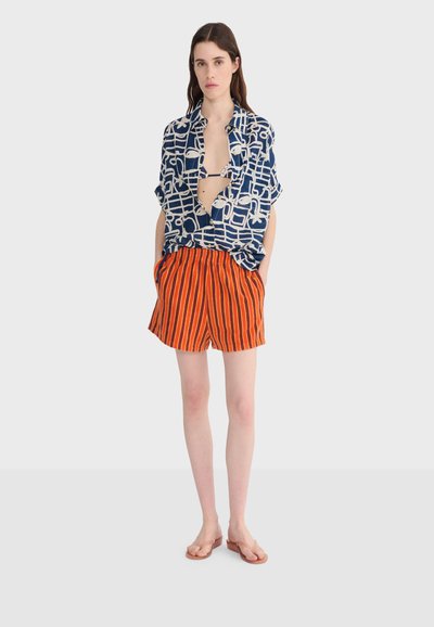 Femme portant une chemise ample à motifs bleu marine et blanc par-dessus un haut de bikini, un short rayé orange et noir, et des sandales marron.