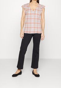 Blouse à carreaux en rose clair, orange et blanc avec des manches courtes et un col en V. Associée à un pantalon noir ajusté et des chaussures plates noires.