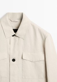 Beiges Langarmhemd mit spitzem Kragen, durchgehender Knopfleiste und zwei Klappentaschen auf der Brust vor weißem Hintergrund.