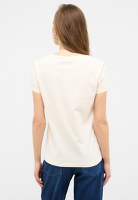 Cremefarbenes T-Shirt mit kurzen Ärmeln, aus Baumwolle, mit Rundhalsausschnitt und leicht abgerundetem Saum, kombiniert mit dunkelblauen Jeans.