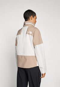 Let léger jakke med et beige og hvidt farveblok design, med et struktureret ternet mønster og høj krave. Logo på ryggen.