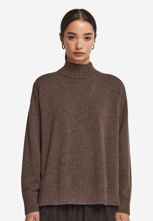 Femme aux cheveux foncés portant des boucles d'oreilles en or en forme d'anneau et un pull à col roulé brun ample, sur un fond blanc uni.