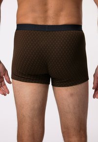 Boxers marron à motif avec une ceinture sombre portant le logo. Le matériau semble lisse, avec un design géométrique sur l'ensemble.