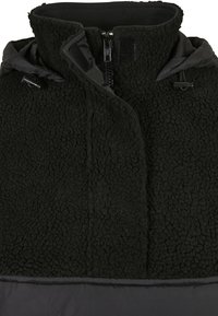 Chaleco de forro polar negro con superficie texturizada y parte inferior de nailon negro lisa. Cuenta con cuello alto, cremallera frontal y cordón ajustable.