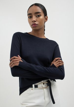 Pull en maille bleu marine à manches longues, col rond et poignets côtelés. Assorti avec un jean blanc et une ceinture noire avec une boucle en or.