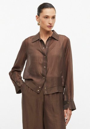 Femme portant un chemisier marron transparent à manches longues boutonné et un pantalon large assorti, cheveux tirés en arrière et boucles d'oreilles créoles dorées.