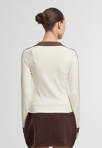 Kerman pitkähihaine paita, jossa on ruskea kaulus ja yksityiskohtia, muotoiltu malli ja sileä pinta. Olkapäillä näkyvät tikkaukset.