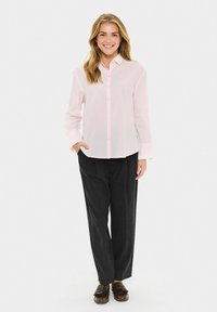 Femme souriante portant une chemise rose clair à boutons, un pantalon noir ample et des mocassins marron, se tenant avec une main dans sa poche.