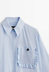 Chemise bleu clair manches longues avec col pointu et poche avant ornée de bords froncés et d’un bouton noir décoratif.