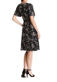 Vestido floral con mangas cortas, tejido negro con flores rosas, cintura ajustada y falda acampanada. Llevado con zapatos de tacón altos negros.