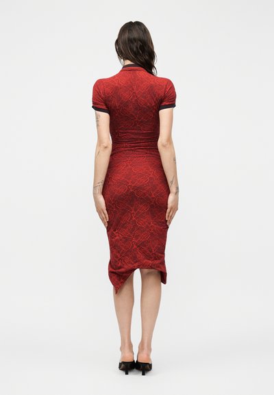 Γυναίκα με μακριά σκούρα μαλλιά που φοράει εφαρμοστό κόκκινο φόρεμα μέσης μήκους με σχέδιο και μαύρα τακούνια, στέκεται με την πλάτη προς τα εμάς σε λευκό απλό φόντο.