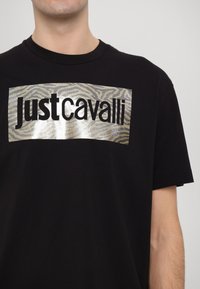 Juodas medvilnės marškinėliai su metaliniu sidabriniu zebro raštu keturkampiu grafiniu elementu, ant kurio drąsiai užrašyta "Justcavalli" juoda spalva.