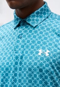 Κοντινό πλάνο ενός ατόμου που φοράει μπλε πόλο πουκάμισο με μικρά λευκά σχέδια καβουριών και λευκό λογότυπο Under Armour στο στήθος.