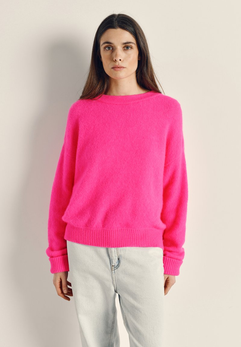 Donna con capelli lunghi castani che indossa un maglione rosa brillante e jeans azzurri, in piedi su uno sfondo chiaro e uniforme.