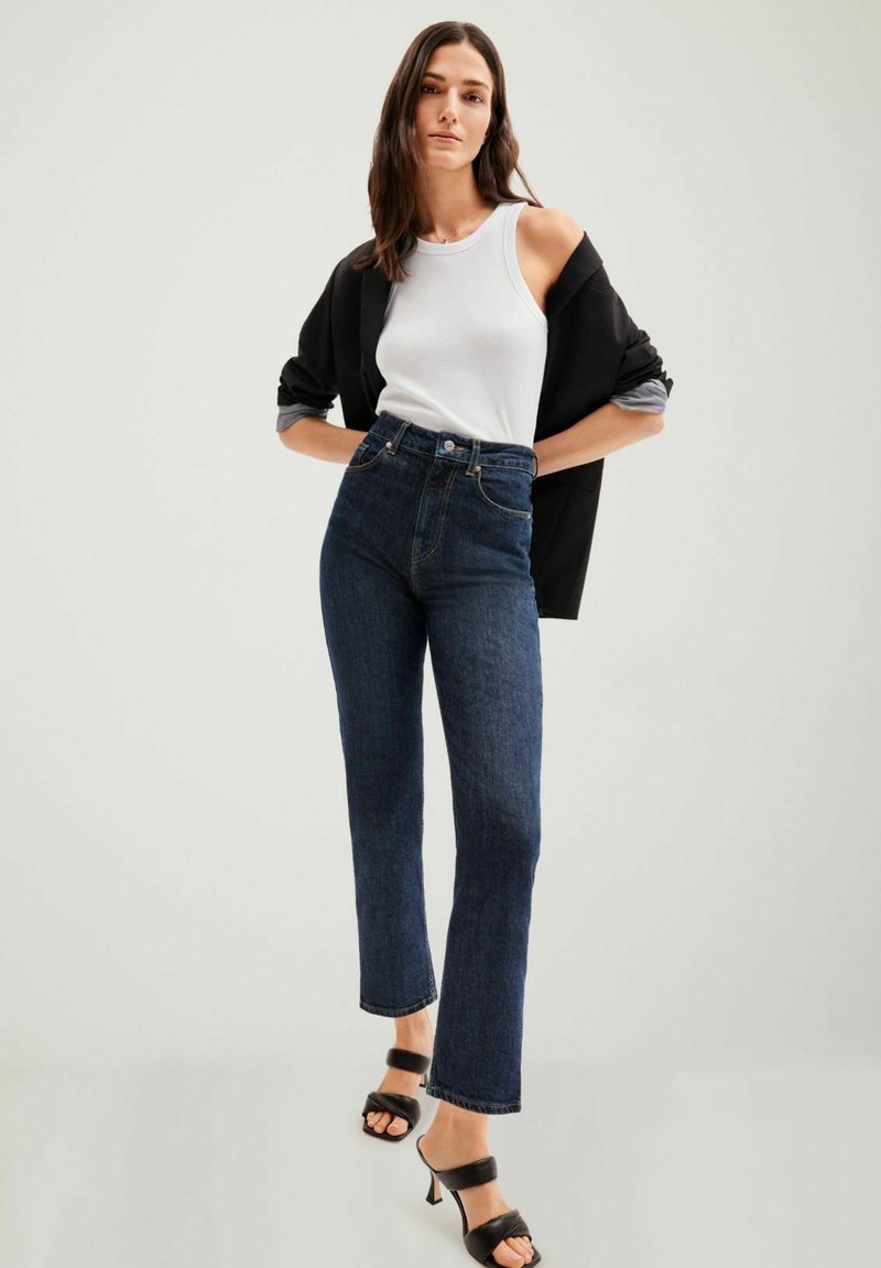 Jean noir taille haute avec une jambe droite, associé à un débardeur blanc et une veste noire oversize, ainsi qu'à des sandales à talons noires.