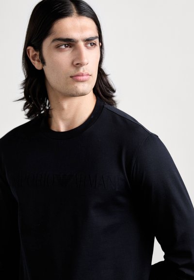 Svart langermet skjorte laget av glatt stoff, med en diskret hevet "Emporio Armani"-logo over brystet og rund hals.
