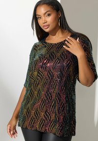 Top nero con paillettes e motivo a onde multicolore, maniche corte, scollatura tonda e vestibilità ampia. Indossato sopra leggings neri.