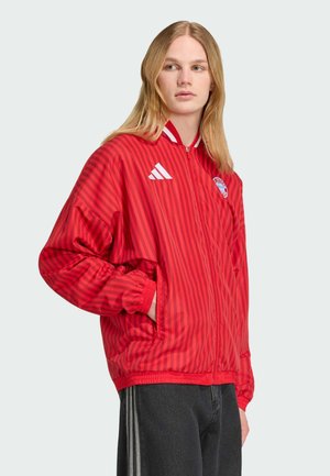 Młoda osoba o długich blond włosach, ubrana w czerwoną kurtkę zapinaną na zamek z paskami i logo Adidas oraz Bayern Monachium, w połączeniu z ciemnymi spodniami.