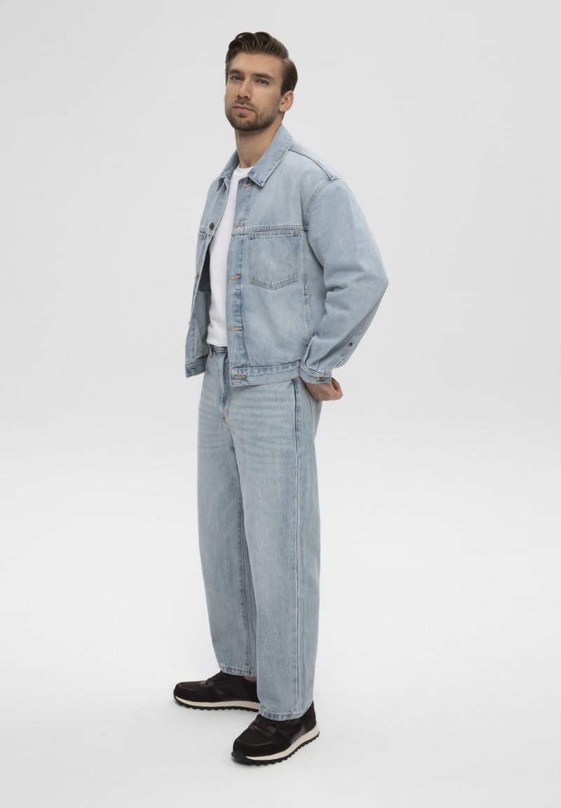 Homme debout, les mains derrière le dos, portant une veste en jean bleu clair, un jean assorti, une chemise blanche et des baskets noires sur fond blanc.