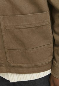 Chaqueta marrón con textura que cuenta con dos grandes bolsillos frontales, contrastada con una camiseta de rayas blanca y beige debajo. Los bordes están cosidos.