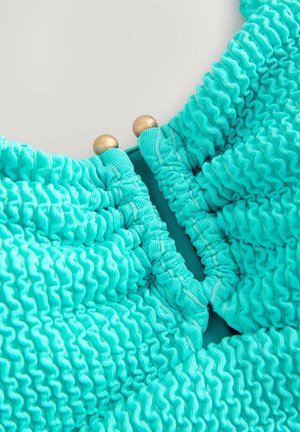 Tissu turquoise froncé présentant une texture froncée, agrémenté de deux boutons décoratifs dorés au niveau du col.