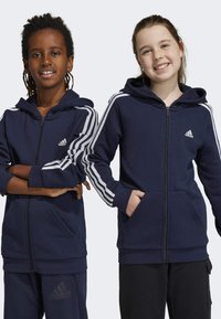 Las sudaderas con cremallera en color navy, de tejido suave, presentan acentos en blanco con tres rayas en las mangas, bolsillos frontales y un pequeño logo en el pecho.