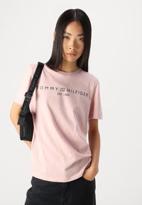Lys rosa bomull t-skjorte med korte ermer, med svart trykt merkenavn "TOMMY HILFIGER EST. 1985" på forsiden. Casual passform.