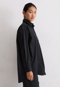 Jonge vrouw in een oversized zwart overhemd met knopen staat zijwaarts tegen een plain witte muur.