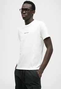 Balts kokvilnas T-krekls ar īsām piedurknēm un apaļu kakla izgriezumu, priekšpusē ar melnu "REPLAY" logotipu. Gluda tekstūra, minimālistisks dizains.