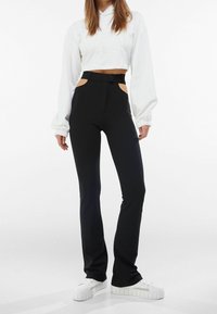 Svarta och figurnära byxor med utskärningar vid midjan, kombinerat med en vit croppad hoodie och vita plattforms-sneakers. Slät känsla.