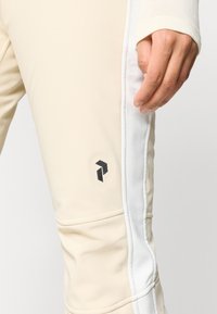 Κρεμ χρώματος αθλητικού παντελονιού με λευκές ρίγες στο πλάι, διαθέτει ένα μικρό μαύρο λογότυπο στο μηρό και έχει απαλό, ελαστικό υλικό.