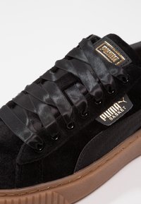 Svart mockaskor med satinsnören, guldlogotagg och sula av gummigummi. Har rund tå och texturerade detaljer på sidorna.