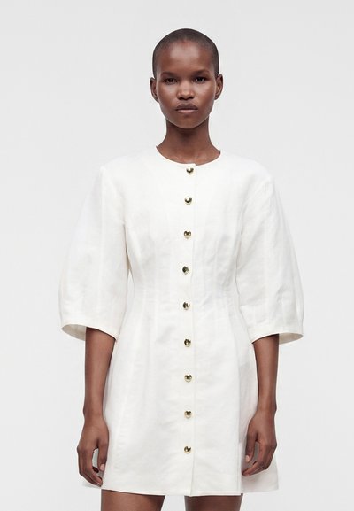 Femme portant une robe blanche à manches longues avec des boutons dorés sur le devant, debout devant un fond clair uni.