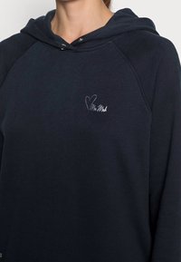 Donna che indossa una felpa con cappuccio blu navy con un piccolo cuore ricamato bianco e testo in corsivo sul petto.
