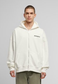 Hvid zip-up hoodie lavet af blødt stof, med en kængurulomme, broderet logo på brystet og en afslappet pasform med hætte.