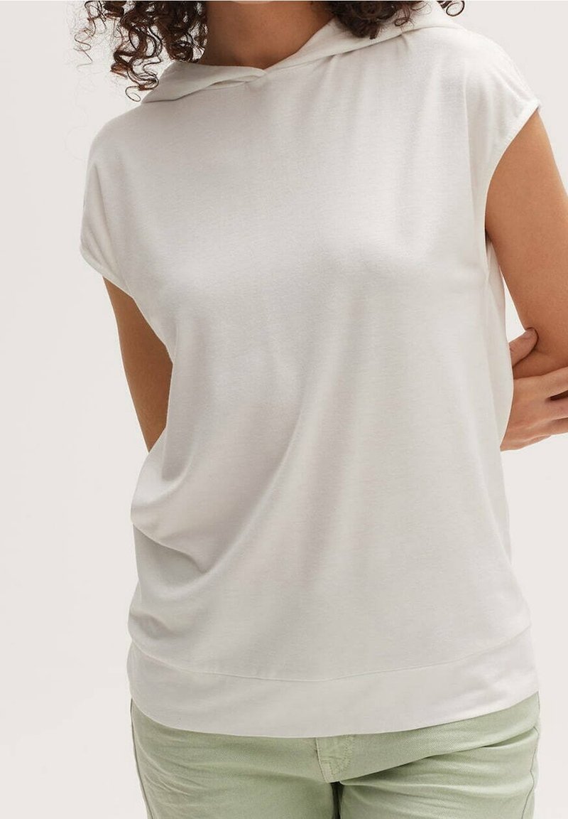 Personne aux cheveux bouclés portant un haut sans manches à capuche de couleur beige clair et un pantalon vert clair, debout avec un bras plié derrière le dos.