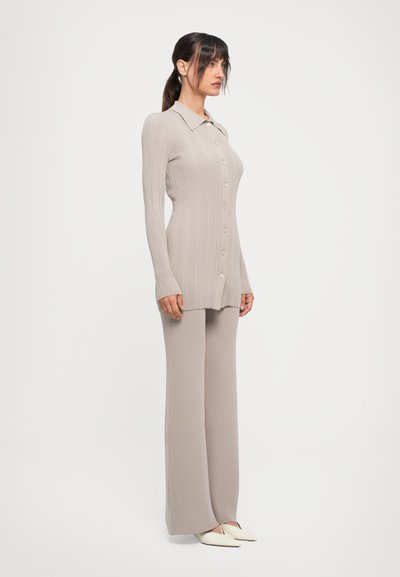 Woman with dark hair in a low ponytail wearing a beige ribbed buttoned cardigan and matching wide-legged pants with cream pointed shoes.