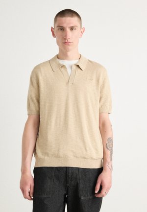 Beige gestricktes Poloshirt mit kurzem Kragen und kurzen Ärmeln, mit lockerer Passform und strukturierter Oberfläche. Getragen mit dunklen Jeanshosen.