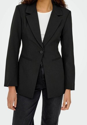 Frau trägt einen taillierten schwarzen Blazer über einem weißen Top und dunklen Jeans, steht mit locker herabhängenden Armen an den Seiten.