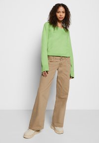 Sweater verde de malha com mangas largas, combinado com calças largas bege e sapatos robustos de cor clara. Fundo simples.