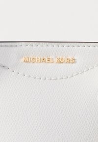 Biała portmonetka z fakturowanej skóry, zdobiona złotym wytłoczonym logo "MICHAEL KORS" na górze. Gładkie przeszycia wzdłuż krawędzi dodają szczegółu.