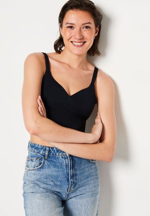 Jonge vrouw met kort donker haar, gekleed in een zwarte mouwloze top en blauwe jeans, staat met gekruiste armen en een glimlach.