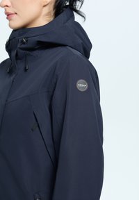 Giacca impermeabile blu navy con tasca con zip, cappuccio regolabile e logo ricamato sulla manica. Texture liscia e design moderno.
