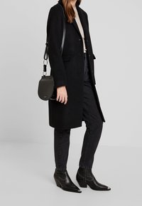 Frau trägt einen schwarzen knielangen Mantel, schwarze Jeans, schwarze spitze Stiefel mit silbernen Spitzen und trägt eine schwarze Lederschultertasche.