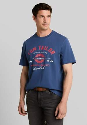 Man draagt een blauw Tom Tailor T-shirt met rode en witte tekst, donkere spijkerbroek en een bruine riem, kijkt iets naar links.