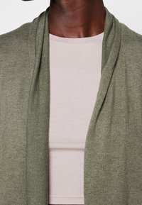 Oliivivihreä neuletakki pehmeällä tekstuurilla, avointa etuosaa, kerrostettuna vaaleanpunaisen ribbipaidan päälle, mikä tuo esiin hienovaraisen värikontrastin.